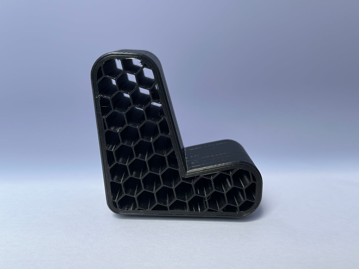 Un Porta telefono nero, a forma di L, stampato in 3D di Il mio negozio con una struttura interna a nido d'ape visibile è incastonato su uno sfondo azzurro sfumato. L'oggetto dal design futuristico ha i bordi arrotondati, appare cavo all'interno e rimane stabile grazie alla sua costruzione innovativa.