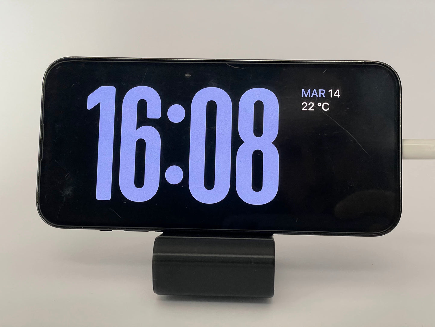 Uno smartphone in orientamento orizzontale visualizza un orologio digitale che indica 16:08. La data "Mar 14" e la temperatura "22°C" sono nell'angolo in alto a destra. Il telefono è montato su un supporto nero con un cavo di ricarica collegato sul lato destro. Questo Porta telefono de Il mio negozio vanta un design accattivante e stabile non scivola.